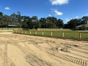 A wooden post-and-rail fence surrounds a grassy paddock with sandy ground and vehicle tire tracks in the foreground.
