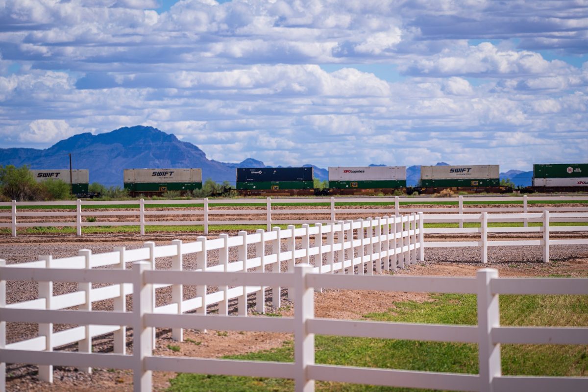 A white ranch-style fence runs across a field with a freight train and mountains in the background.